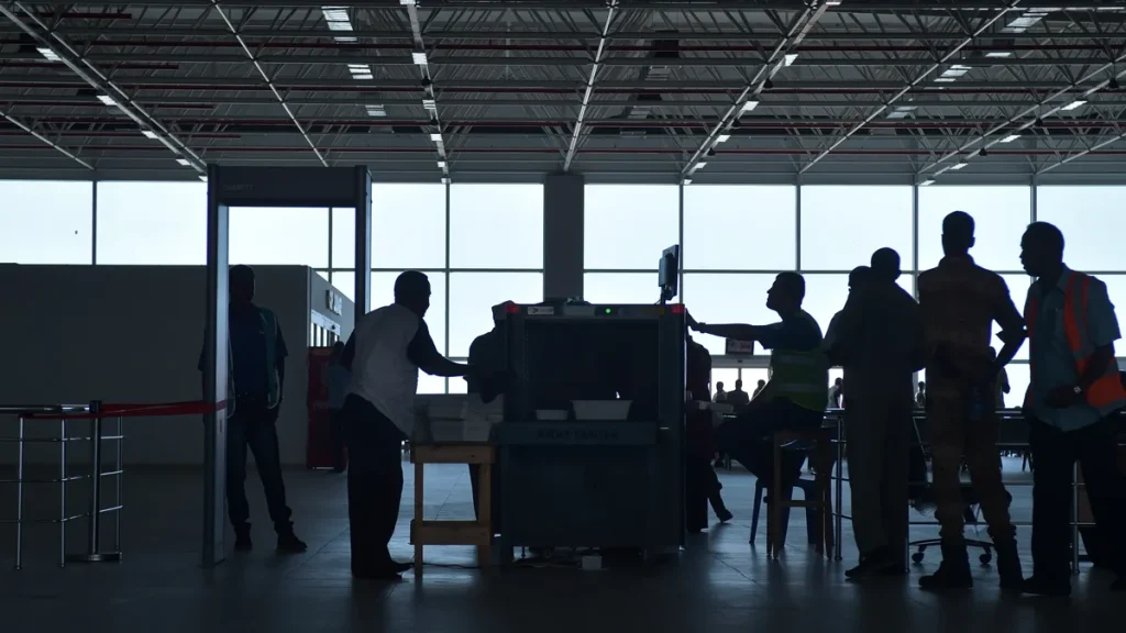 Silhouetted travellers and screeners at an airport X-ray baggage scanner and walk-through metal detector
